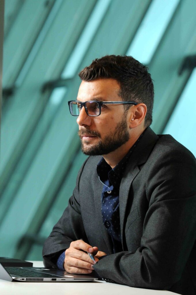 Rodrigo De Vecchi, wearing a dark blazer, hands clasped, mid-discussion.